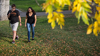 A male and female student walking on campus in the early autumn. Leaves can be seen on the ground and in the forefront of the picture.